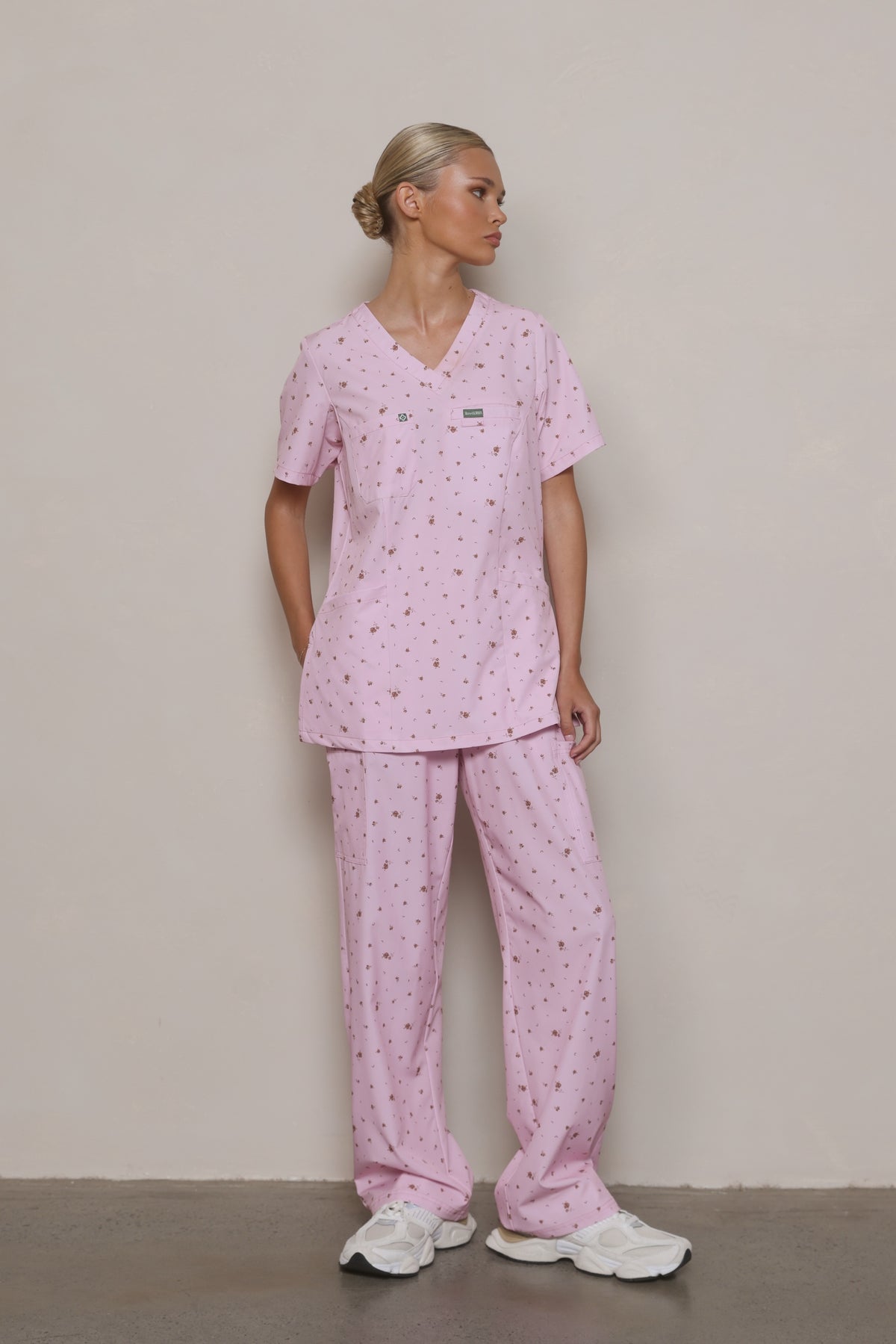 Model wearing a Hero Scrubs pastel pink floral scrub set with one hand in her pocket, looking to the side.