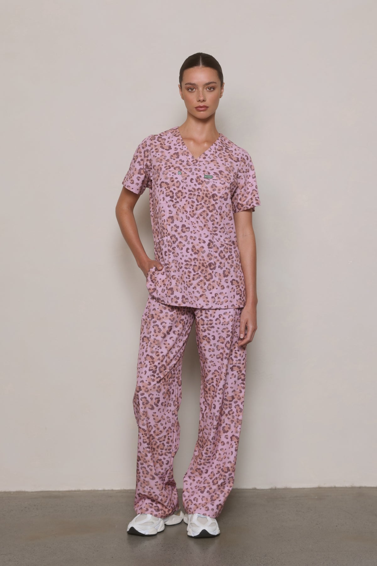 Length View Full-length view of a model wearing a Hero Scrubs pink leopard-print scrub set with one hand on her hip.