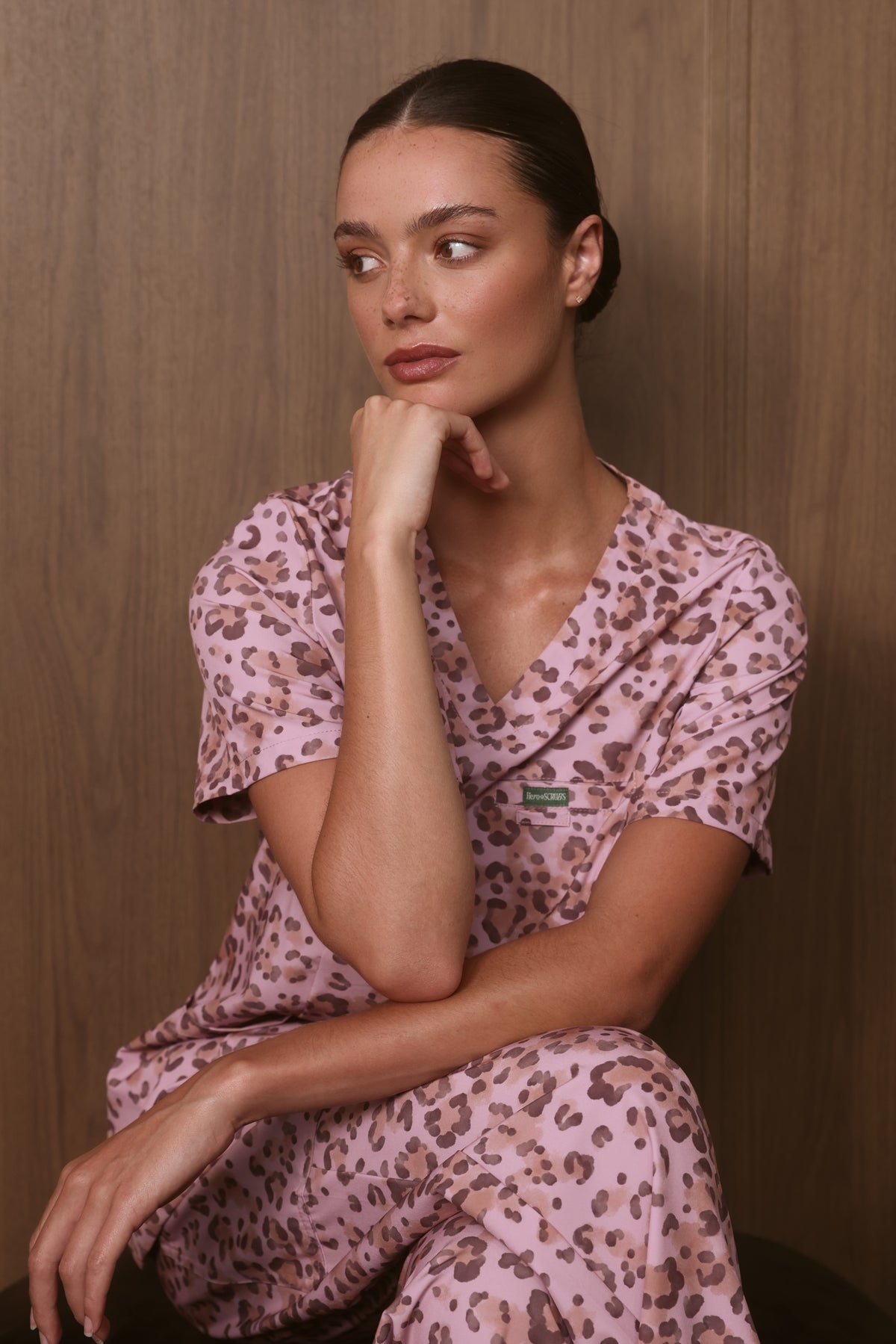 Seated model wearing a Hero Scrubs pink leopard-print scrub set against a timber backdrop.