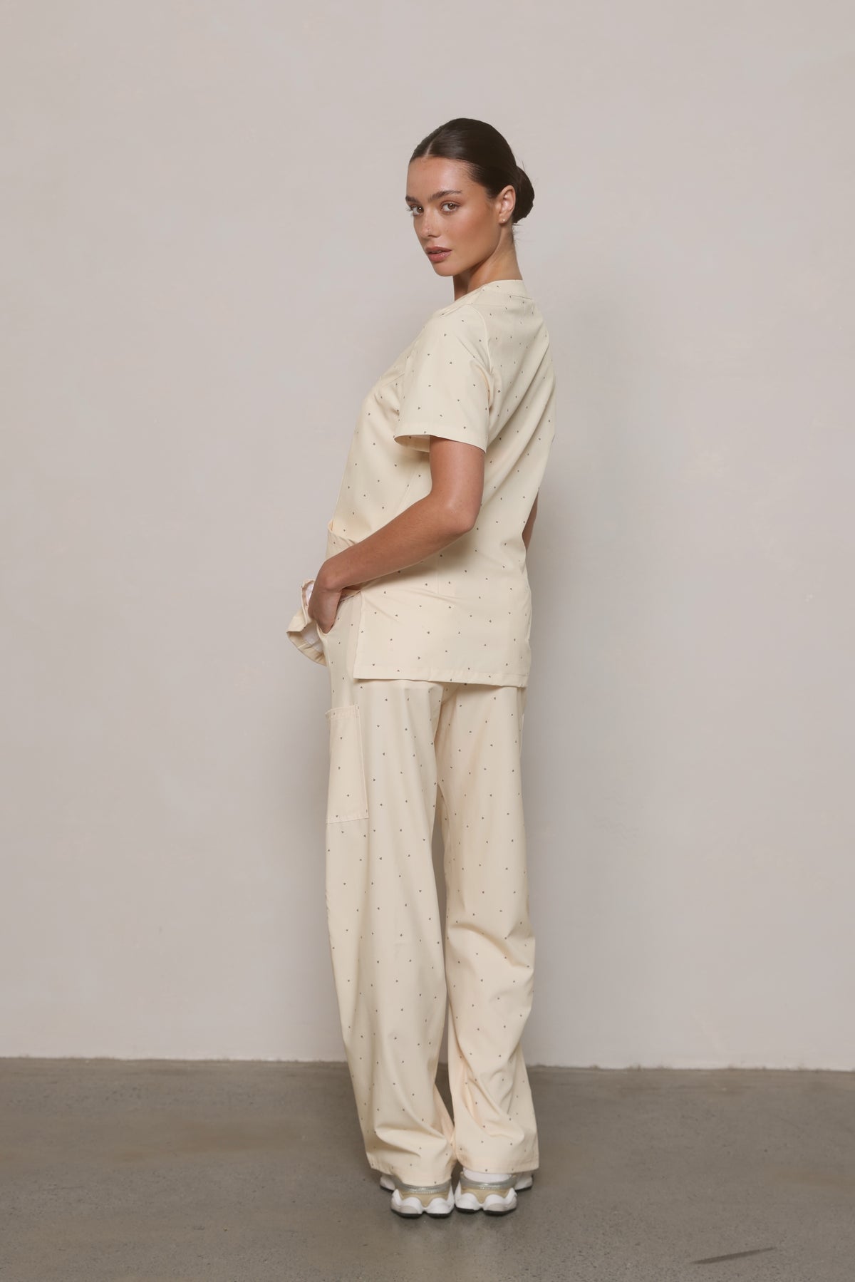 Model wearing a Hero Scrubs cream printed scrub set, turned to show the side of the top and pants.